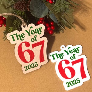 K&ouml;nnte beinhalten: Zwei Holzornamente mit dem Text "The Year of 67 2025" in Gr&uuml;n und Rot. Ein Ornament befindet sich auf einem nat&uuml;rlichen Holzgrund, das andere ist wei&szlig;. Die Ornamente befinden sich in der N&auml;he einer dekorativen Anordnung aus Gr&uuml;n und roten Beeren.