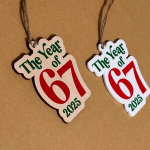 K&ouml;nnte beinhalten: Zwei Holzornamente mit dem Text "The Year of 67 2025" in Gr&uuml;n und Rot. Eines ist aus Naturholz, das andere wei&szlig;. Beide haben eine Schnur zum Aufh&auml;ngen.