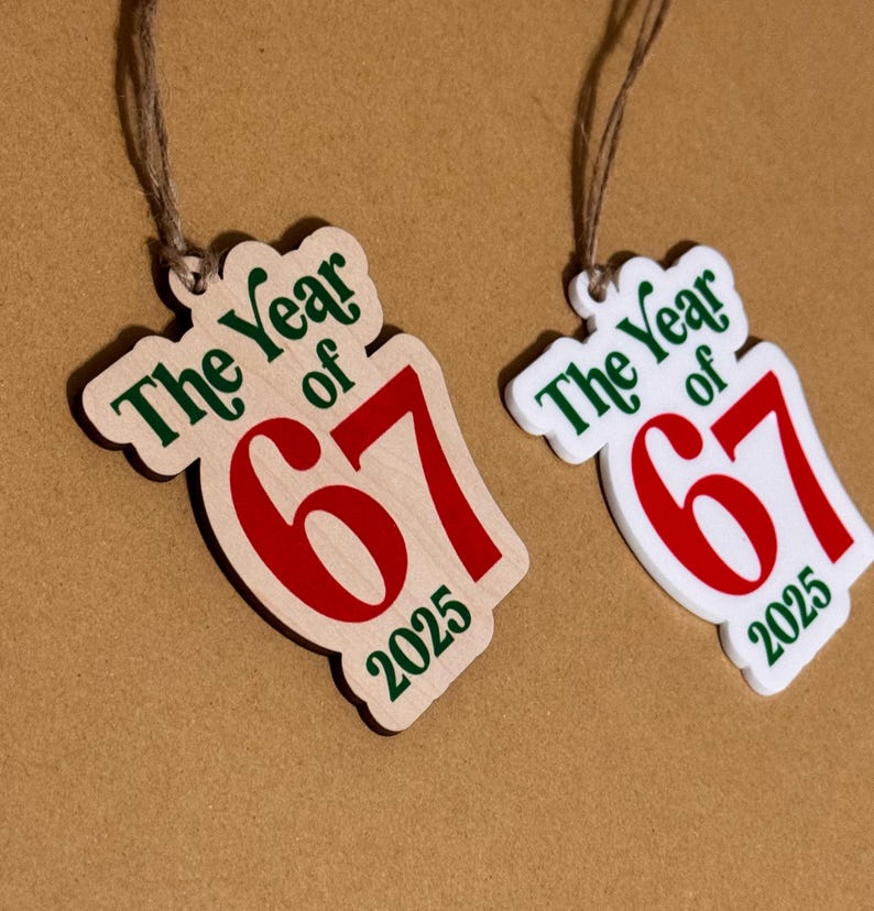 K&ouml;nnte beinhalten: Zwei Holzornamente mit dem Text "The Year of 67 2025" in Gr&uuml;n und Rot. Eines ist aus Naturholz, das andere wei&szlig;. Beide haben eine Schnur zum Aufh&auml;ngen.