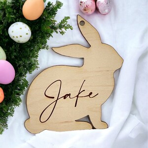 May include: A wooden bunny rabbit cutout with the name "Jake" engraved on it. The bunny is light brown and is surrounded by pastel colored Easter eggs.