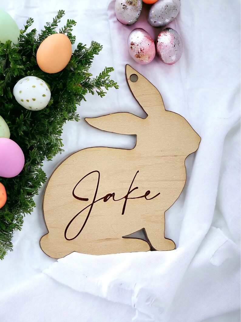 May include: A wooden bunny rabbit cutout with the name "Jake" engraved on it. The bunny is light brown and is surrounded by pastel colored Easter eggs.