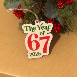 K&ouml;nnte beinhalten: Weihnachtsschmuck aus Holz mit dem Text "The Year of 67 2025" in Gr&uuml;n und Rot. Der Schmuck h&auml;ngt an einer Schnur und ist von Gr&uuml;n und roten Beeren umgeben.