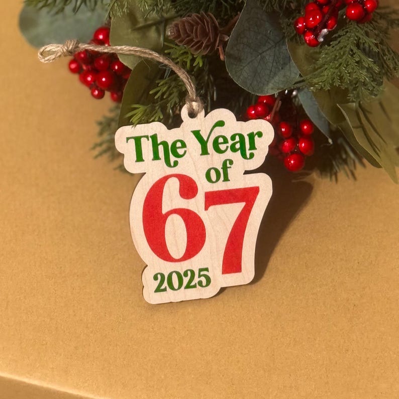 K&ouml;nnte beinhalten: Weihnachtsschmuck aus Holz mit dem Text "The Year of 67 2025" in Gr&uuml;n und Rot. Der Schmuck h&auml;ngt an einer Schnur und ist von Gr&uuml;n und roten Beeren umgeben.