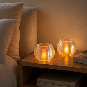 May include: Two clear, ribbed glass candle holders sit on a wooden nightstand. Each holder contains a glowing, warm-toned light source. Below the holders, two stacked books are visible, with the top book titled A Democracy of Imagery.