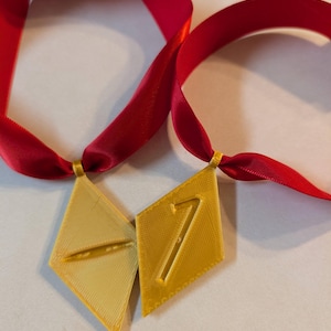 May include: Two gold-colored diamond-shaped medals with the numbers 7 and a horizontal line, attached to red satin ribbons. The medals are likely awards or recognition items, suitable for events or achievements.