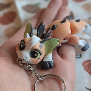 May include: A 3D-printed articulated dragon keychain featuring a peach-colored body with white accents, black paws, and light green wing details. The creature has large black eyes with white highlights and a silver metal split ring attached.