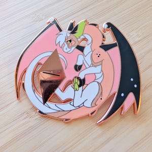 May include: A rose gold enamel pin featuring a stylized dragon with white, peach, and black accents. The dragon has a white mane with green highlights and is set against a pink background with black wings.