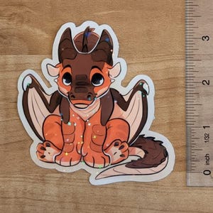 Pu&ograve; includere: Un adesivo olografico di un drago dei cartoni animati con squame marroni e arancioni, grandi occhi e ali, posizionato accanto a un righello che mostra che l'adesivo &egrave; alto circa 7,6 cm. Il drago ha il ventre chiaro e una lunga coda.
