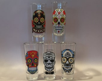 May include: Set of five clear glass shot glasses with colorful sugar skull designs. The designs include a skull with a cross, a skull with a bow tie, a skull with flowers, and two skulls with flowers and text.