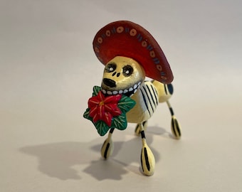 May include: A yellow and black skeletal dog figurine wearing a red sombrero and holding a red flower.