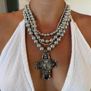 Puede incluir: Un collar plateado de varias capas con un gran colgante de cruz. El collar presenta múltiples hilos de cuentas de diferentes tamaños, que culminan en un colgante de cruz detallado con un corazón en el centro. La cruz es ornamentada con detalles intrincados.