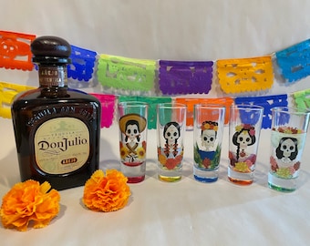 May include: Five colorful shot glasses with Day of the Dead sugar skull designs. The glasses are arranged in a row on a white surface. A bottle of Don Julio Añejo tequila is in the background.