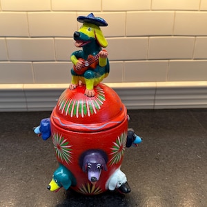 May include: A red ceramic jar with a colorful painted design featuring dogs. The lid is topped with a small figurine of a dog wearing a blue sombrero and playing a guitar.