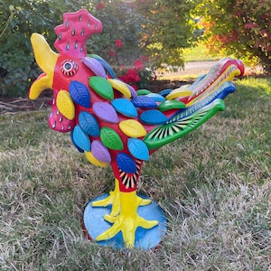 May include: A colorful ceramic rooster figurine with red, yellow, green, blue, and purple leaves painted on its body. The rooster is standing on a blue base with yellow feet.