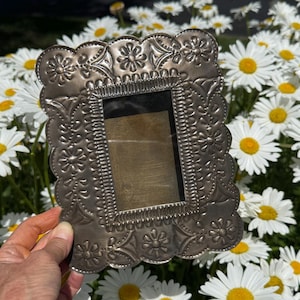 May include: A rectangular metal picture frame with a scalloped edge and a decorative embossed floral pattern. The frame features a metallic silver finish with a dark, antiqued patina in the recessed areas, surrounding a central rectangular opening.
