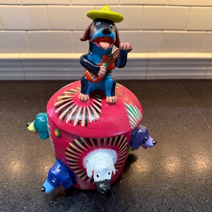 May include: A colorful ceramic piggy bank with a sombrero-wearing dog playing a guitar on top. The piggy bank is decorated with other dogs around the base.
