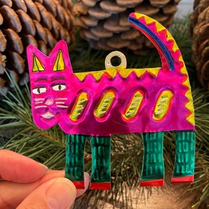 May include: A pink and green metal cat ornament with a yellow and blue tail. The cat has a smiling face with black eyes and a white nose.