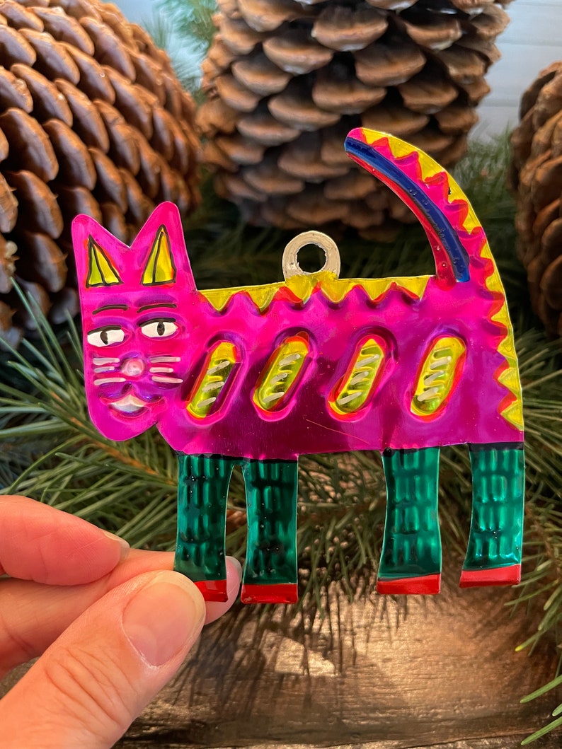 May include: A pink and green metal cat ornament with a yellow and blue tail. The cat has a smiling face with black eyes and a white nose.