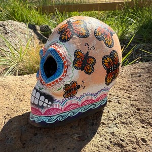 Puede incluir: Una calavera decorativa del Día de Muertos, pintada con mariposas monarca naranjas y negras. La calavera presenta diseños intrincados en azul, rosa y blanco, con un patrón floral detallado en la base. Las cuencas de los ojos son oscuras.