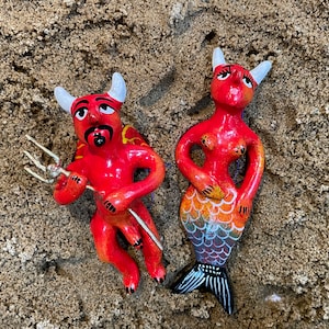 May include: Two red ceramic figurines, one with horns and a trident, the other with a fish tail. Both have black eyes and white horns.