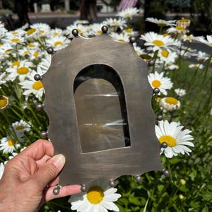 May include: A brushed metal picture frame with a scalloped edge featuring small spherical accents along the perimeter and a central arched opening. The frame has a dark, matte finish with visible horizontal brush strokes across the surface.