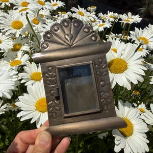 May include: A rectangular bronze-toned metal frame featuring a decorative arched top with a heart motif and embossed floral patterns on the sides. The frame includes a central glass window and a small side latch, presenting a metallic finish.