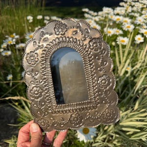 May include: A silver-toned metal picture frame with an arched center opening. The frame features an embossed floral pattern with scalloped edges and a decorative border of small dots surrounding the central aperture for a photograph.