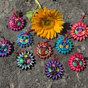 Puede incluir: Nueve adornos circulares con forma de sol, con rostros pintados a mano y detalles florales en colores vibrantes como rosa, azul, naranja y morado, cada uno con una cinta roja para colgar, dispuestos alrededor de un gran girasol amarillo.