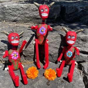 May include: Three red paper mache skeleton figures with painted details, including flowers and bones. The figures are sitting on a rock surface with orange marigold flowers in front of them.