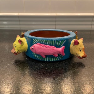 May include: A blue ceramic planter with three yellow animal heads and a pink fish painted on the front. The planter is decorated with green and white stripes.