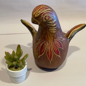May include: A hand-painted ceramic bird figurine with red, yellow and brown accents. The bird has a long beak and outstretched wings. A small potted succulent plant is in the foreground.