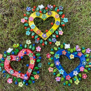 May include: Three heart-shaped wall decorations with a folk art style. Each heart is surrounded by a wreath of colorful flowers, leaves, and butterflies. The hearts are painted in yellow, red, and blue.