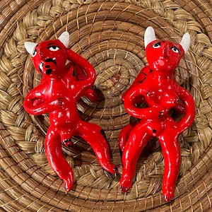 Puede incluir: Dos figuras de cerámica roja de diablos con cuernos y detalles blancos. Las figuras están en posición sentada con las piernas extendidas.