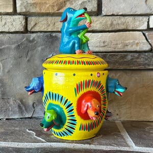 May include: A hand-painted ceramic jar with a yellow base and a lid. The jar features multiple dog figures, including one on top holding a green snake. The jar has a vibrant design with red, blue, and green accents.
