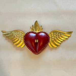 May include: A red heart with gold wings and a flame on top. The heart is decorated with gold studs and has a hinged opening.