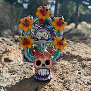 May include: A Day of the Dead-themed decorative skull with a floral headdress. The skull is painted with intricate designs in red, orange, and white. The headdress features orange flowers, teal accents, and small red ornaments.