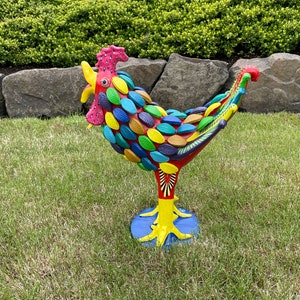 May include: A colorful rooster sculpture made of painted ceramic. The rooster is standing on a blue base and has a red, yellow, and green body with a pink comb and wattle.