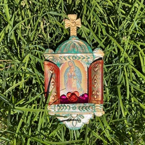 May include: A decorative metal shrine featuring a central image of the Virgin of Guadalupe. The structure has a green domed top with a cross, two hinged red doors with embossed patterns, and a small shelf holding three small red and pink roses.