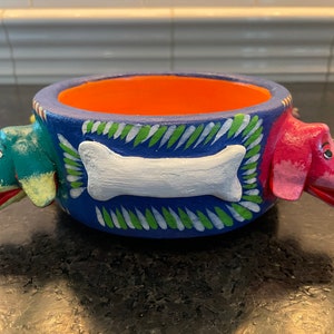 May include: A blue ceramic dog bowl with three dog heads on the rim. The dog heads are painted in green, pink, and yellow. The bowl has a white bone painted on the front.