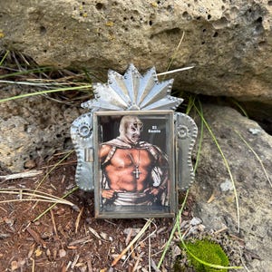 May include: A small, decorative metal frame with a picture of a masked wrestler, El Santo. The frame has a silver finish with a sunburst design at the top. The wrestler is wearing a mask and a cross necklace. The frame is resting on the ground.