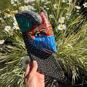 Puede incluir: Una máscara de madera pintada a mano con un hocico puntiagudo, un ojo visible y cuernos curvados. La superficie está decorada con patrones vibrantes que incluyen rayas rojas, blancas y amarillas, una sección azul con puntos y una base negra con motas de colores.