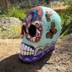 May include: A hand-painted Day of the Dead skull with a light blue background and butterfly accents. The skull features intricate designs in red, white, and black, with a purple base and white teeth. A decorative piece.