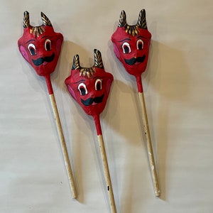 May include: Three red paper mache devil masks on sticks. The masks have black eyes, a black mustache, and gold horns. The sticks are brown.