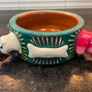 May include: A green ceramic dog bowl with a white bone and three dog faces painted on the side. The dog faces are black, white, and pink.