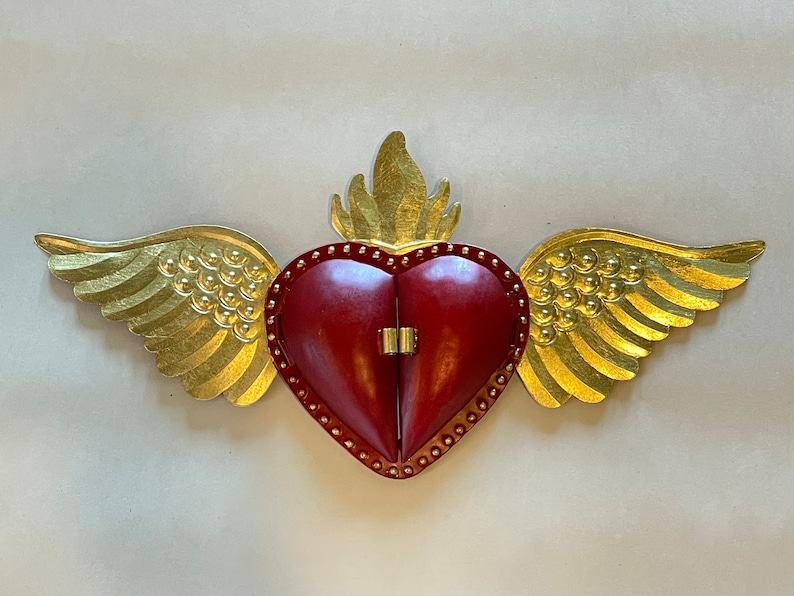 May include: A red metal heart with gold wings and a flame on top. The heart is hinged and opens to reveal an empty space inside.