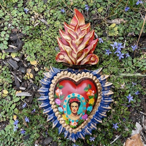 May include: A red heart-shaped wooden ornament with a blue and gold border and a painted image of Frida Kahlo inside. The heart is topped with a carved wooden flame in red and gold.