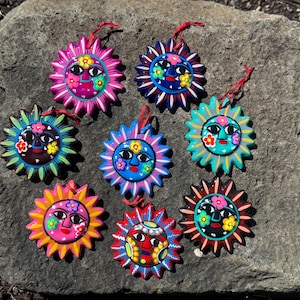 May include: Nine hand-painted sun ornaments in various colours, each with a smiling face and floral details. The suns, in shades of pink, blue, green, and yellow, are strung with a red cord for hanging. Each ornament is unique.