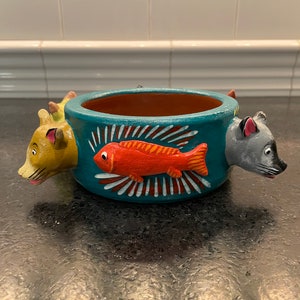 May include: A hand-painted, teal ceramic planter with a red fish design and animal head handles. The planter features a round opening with a brown interior. The handles are a yellow lion and a gray mouse.