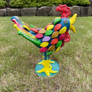 May include: A colorful rooster figurine with a blue body, yellow legs, and a red comb. The rooster is covered in multi-colored leaves and has a bright blue eye.