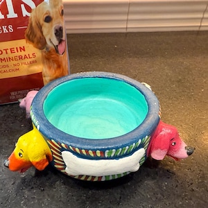 May include: A blue and white ceramic dog bowl with three dog faces around the rim. The bowl has a turquoise blue interior and a white bone painted on the front. The dog faces are yellow, pink, and a light brown.